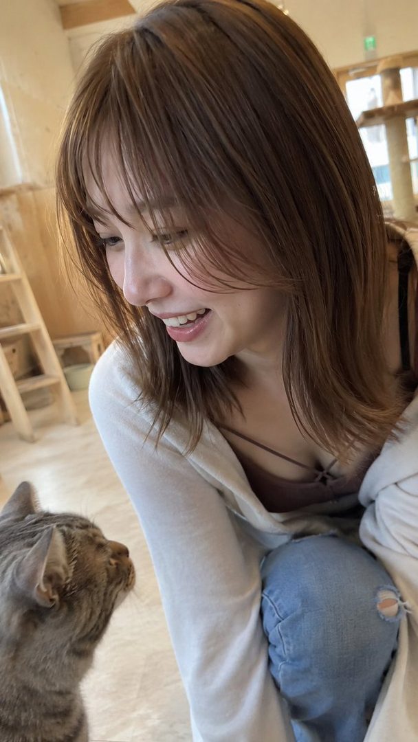 A young woman with brown hair and bangs smiles while looking at a cat in an indoor setting.