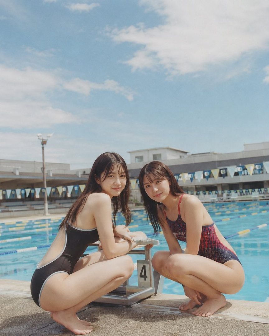Two young women in swimsuits crouch by a swimming pool on a sunny day.