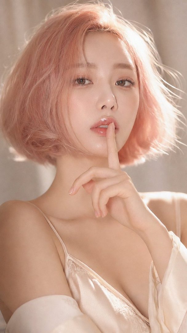 A woman with short pink hair and a light-colored top holds a finger to her glossy lips in a close-up portrait.