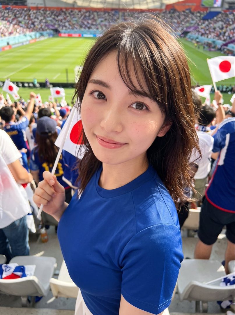 A young woman in a blue jersey holds a Japanese flag at a soccer stadium, smiling at the camera.
