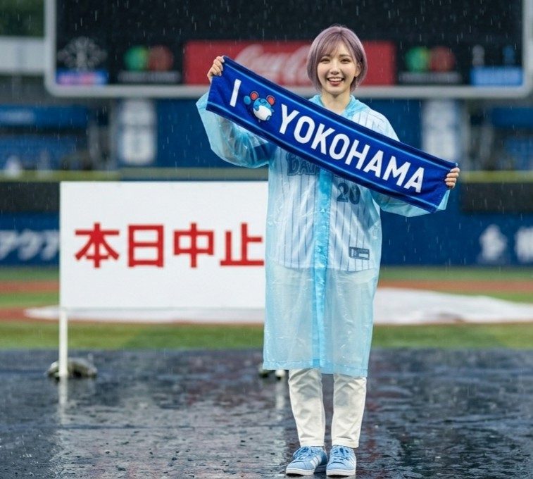 A young woman with short silver hair smiles while holding an "I love Yokohama" towel, wearing a clear raincoat over a baseball jersey on a rainy baseball field.
