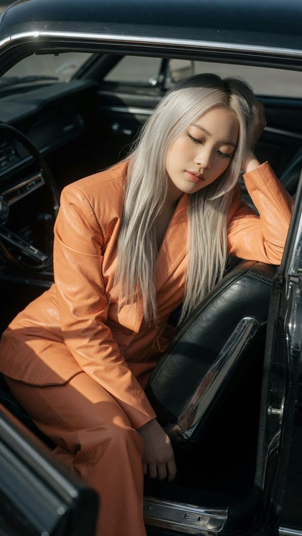 A young East Asian woman with long silver hair and an orange leather suit sits in a vintage black car.