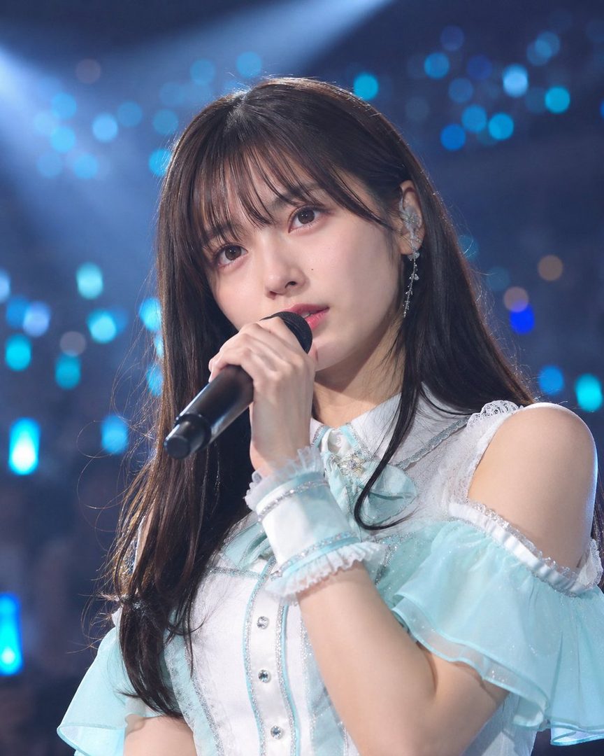A young woman with long dark hair and bangs, wearing a light blue and white dress, sings into a microphone on a stage with blurred blue lights in the background.