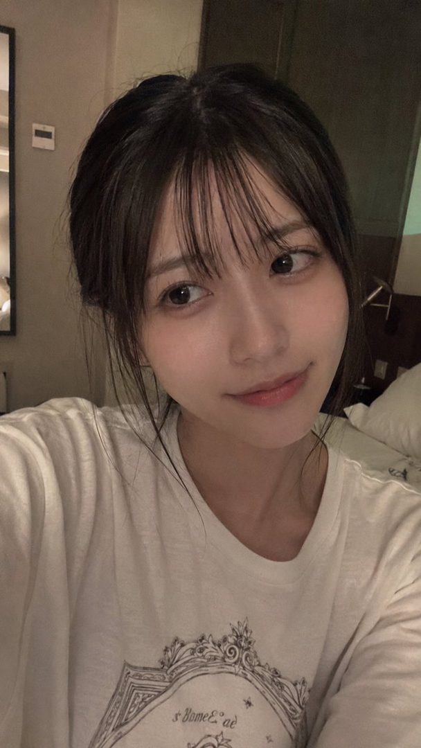 A young woman with dark hair and bangs takes a selfie indoors, wearing a white t-shirt.