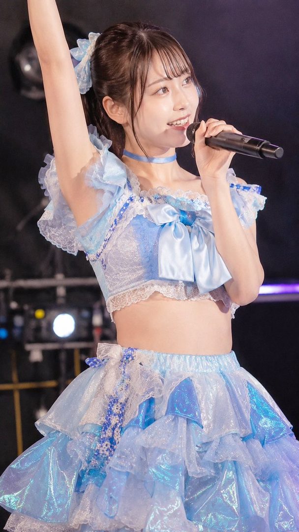 A Japanese idol in a light blue ruffled costume with a microphone, performing on stage.