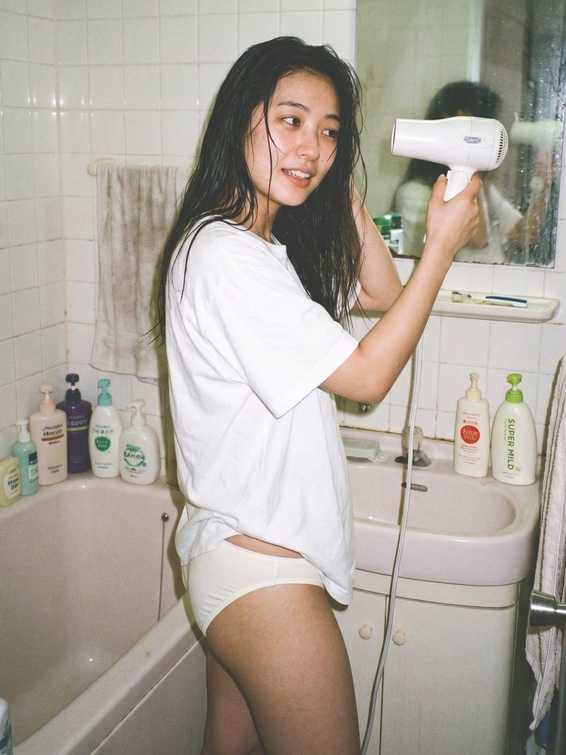 A young woman with wet black hair, wearing a white t-shirt and white underwear, is drying her hair with a hairdryer in a bathroom.