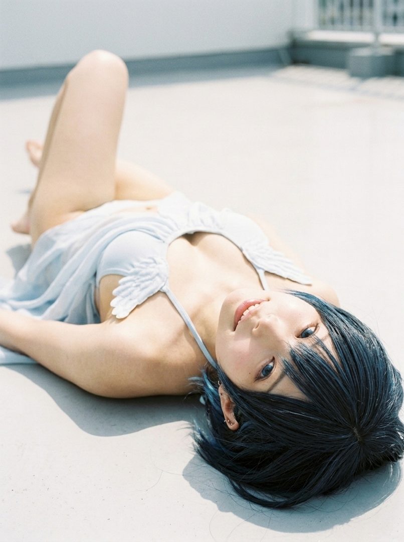 A woman with blue hair and a light blue top lies on her back outdoors, looking up at the camera with a gentle smile.