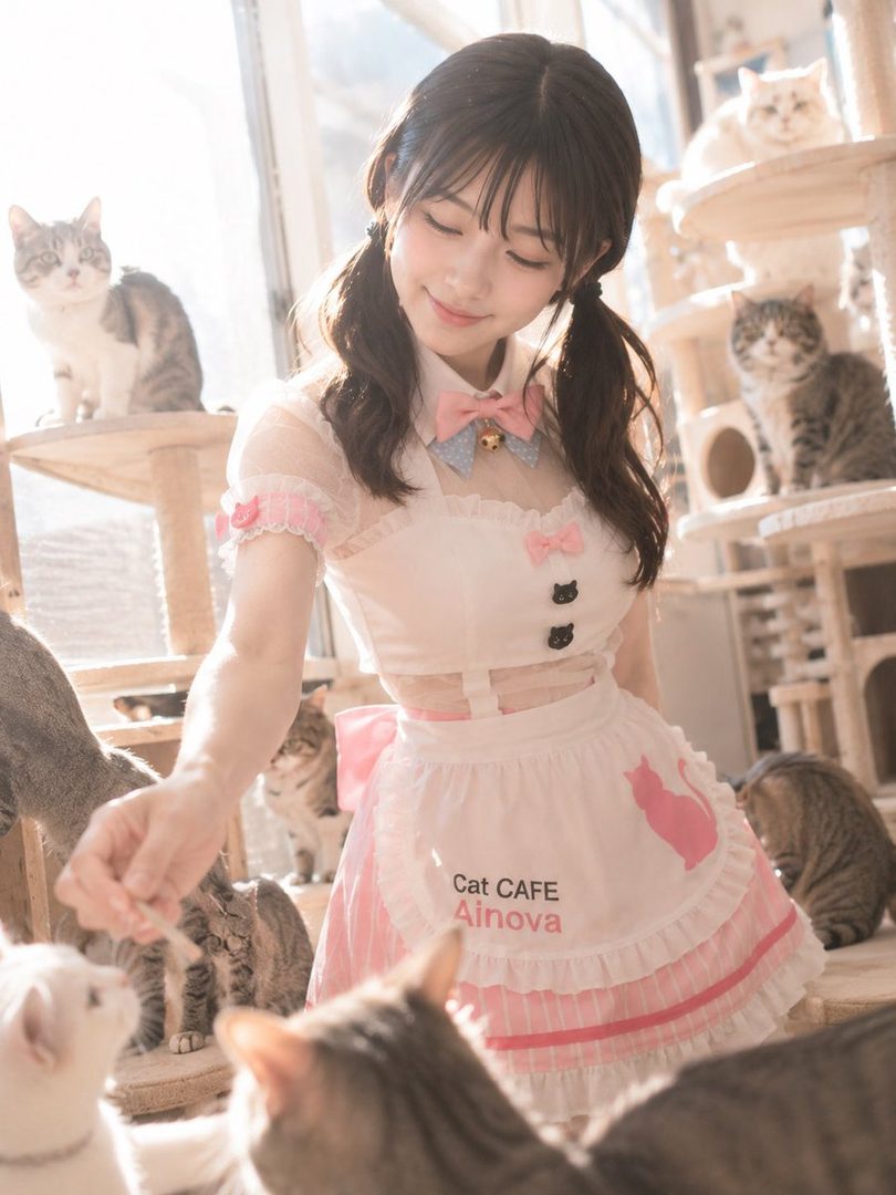 A young woman with brown twintails and bangs, wearing a pink and white maid outfit, smiles softly while feeding cats in a bright cat cafe.