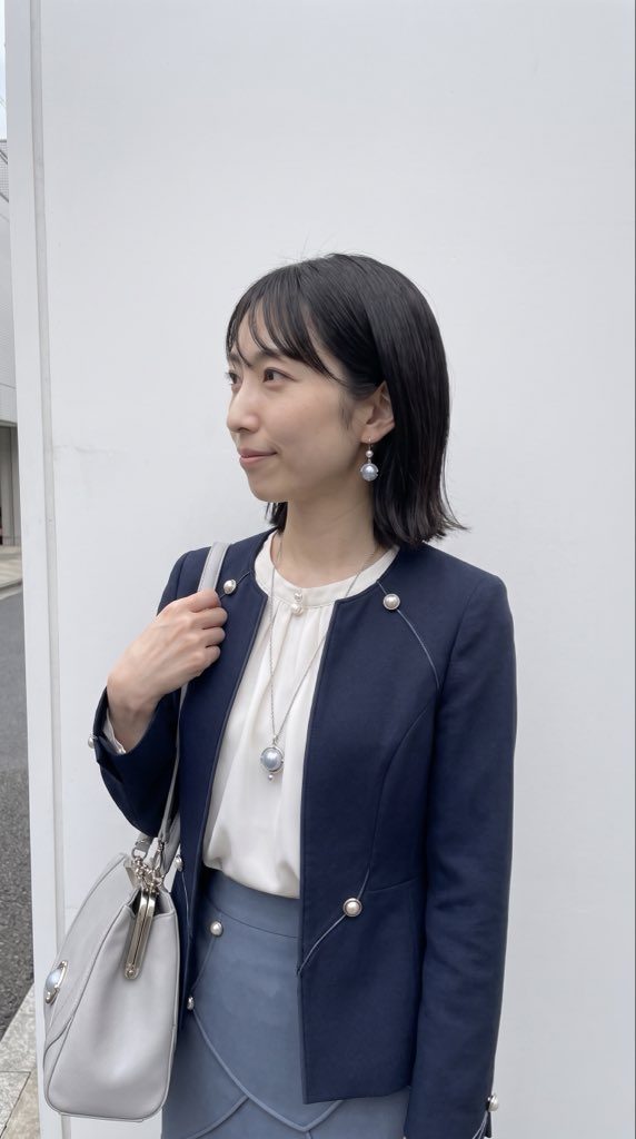 A woman with short black hair and bangs, wearing a navy jacket and a light blue skirt, stands outdoors with a white bag.