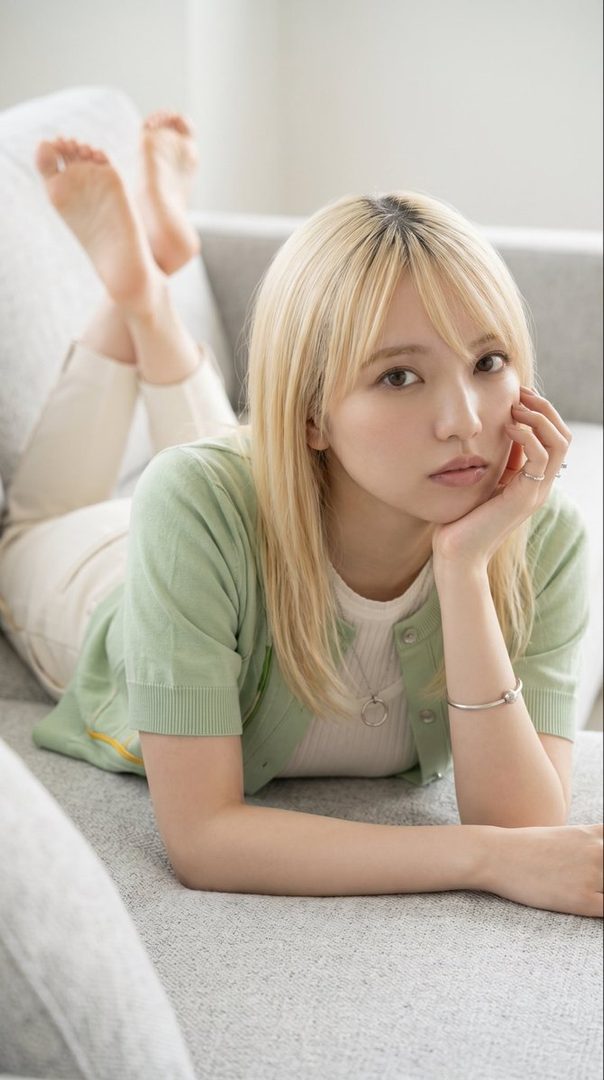 A young woman with blonde hair and bangs, wearing a light green cardigan and white top, lies on a gray sofa indoors, looking calmly at the camera.