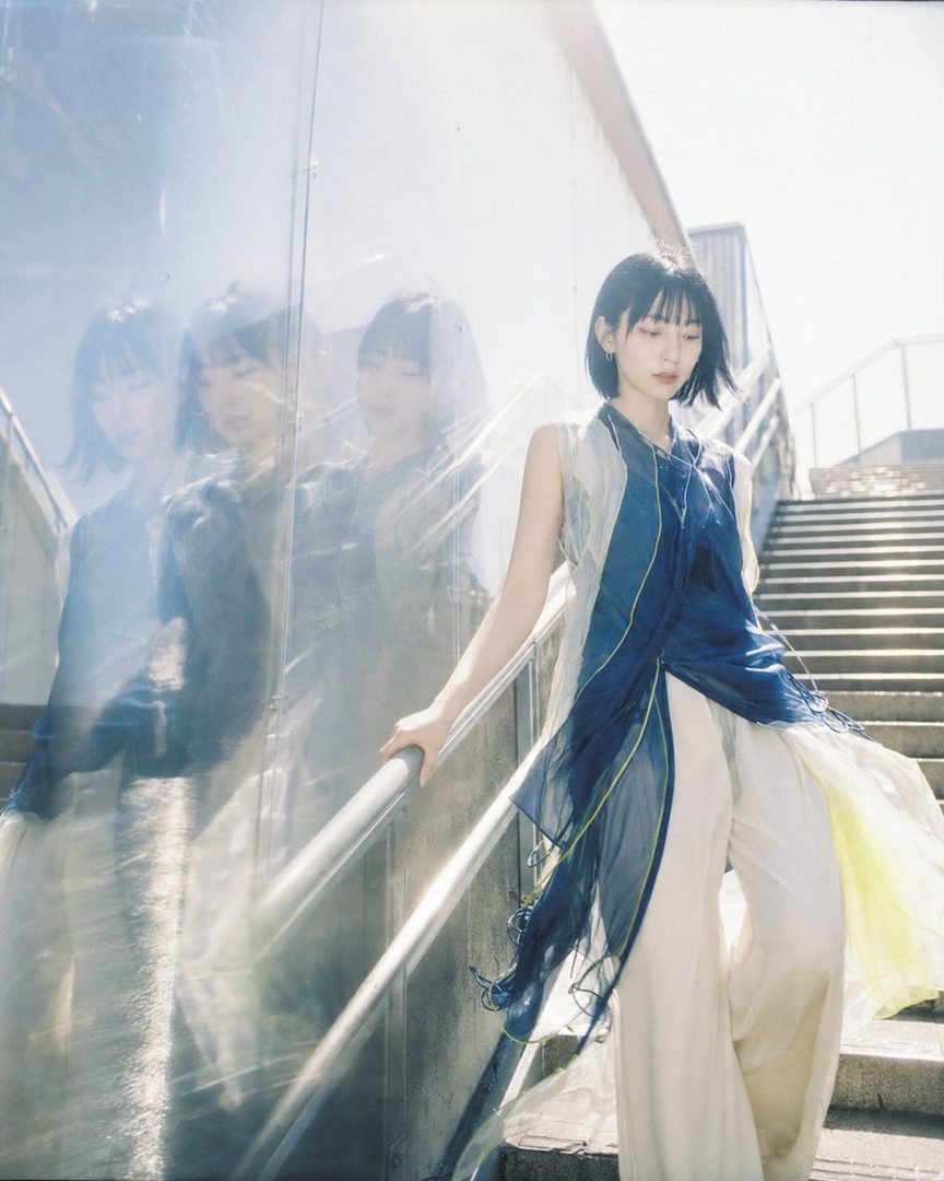 A woman with short black hair wearing a flowing blue and white outfit stands on outdoor stairs, with reflections visible on a glass wall.