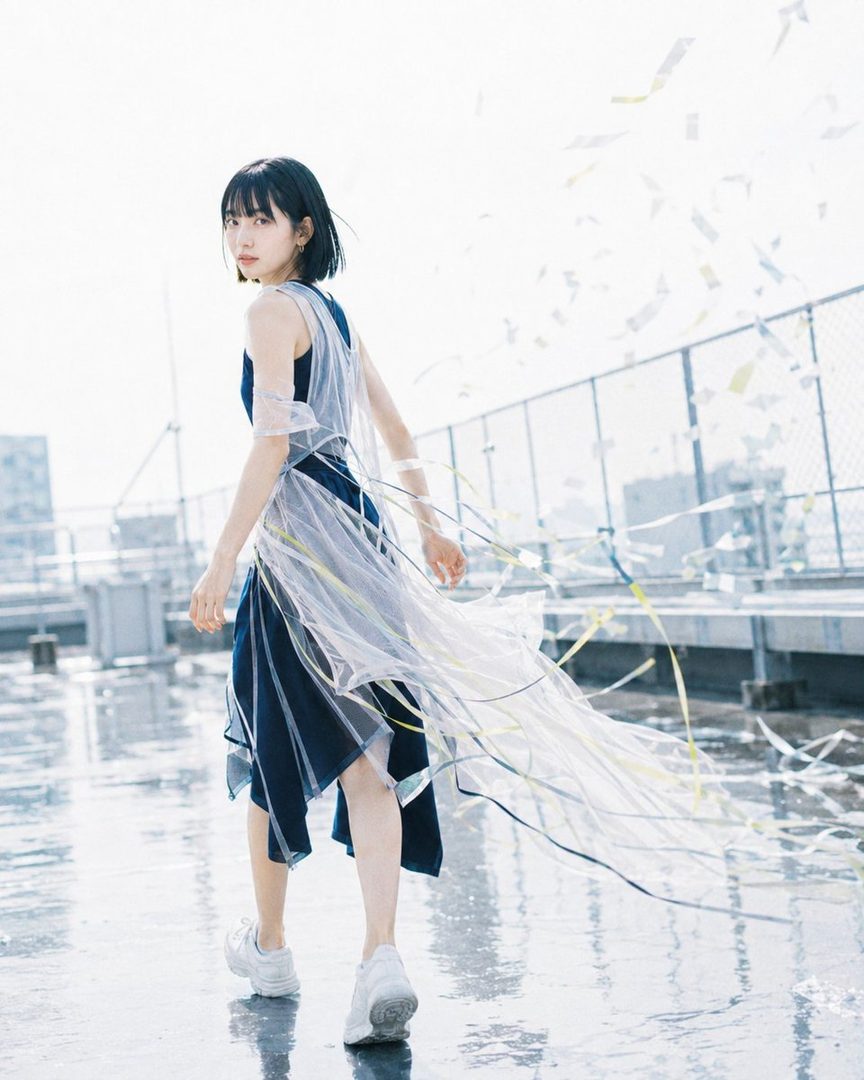 A young woman with short black hair and a blue dress with a sheer white overlay walks on a wet rooftop with confetti in the air.