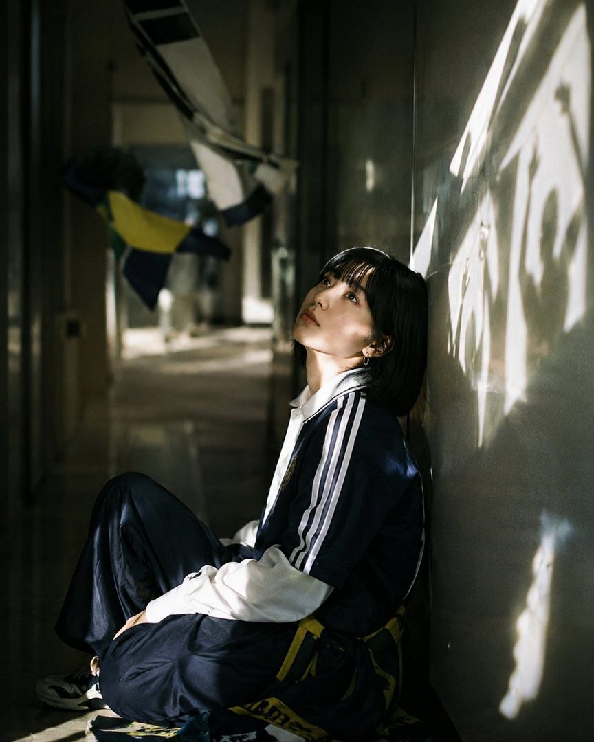 A young woman with short black hair and bangs, wearing a dark blue and white tracksuit, sits on the floor in a dimly lit indoor setting with sunlight casting shadows on the wall.