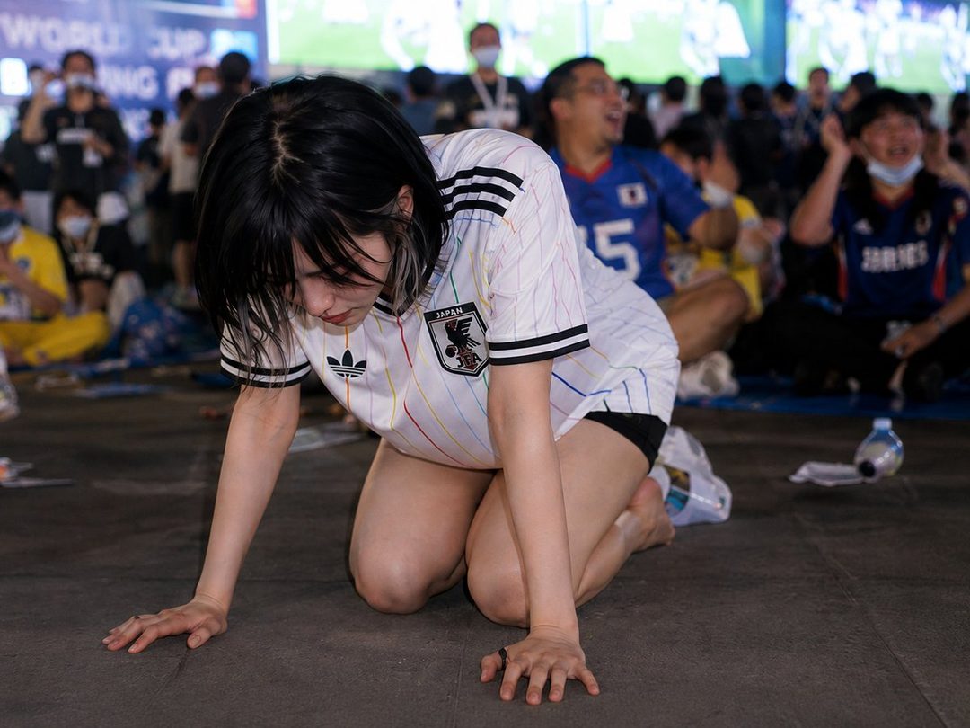 A young woman in a Japan national football team jersey kneels on the ground, looking downcast, surrounded by other spectators.