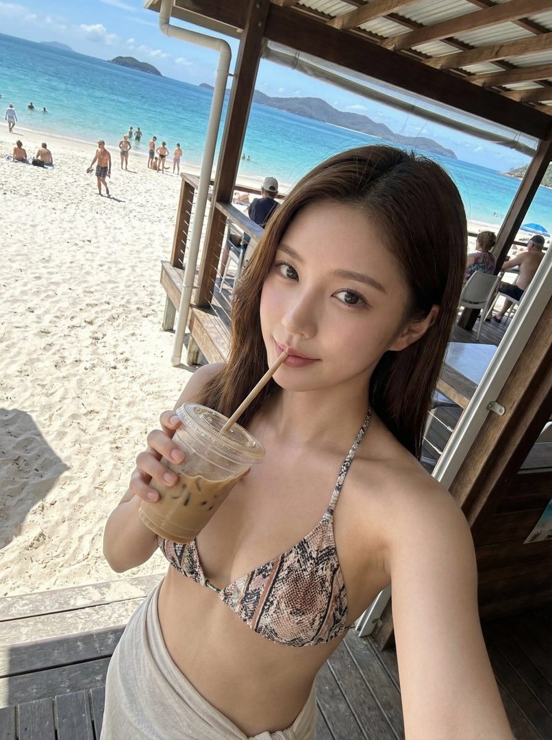 A young woman in a snakeskin print bikini top sips a drink on a sunny beach.