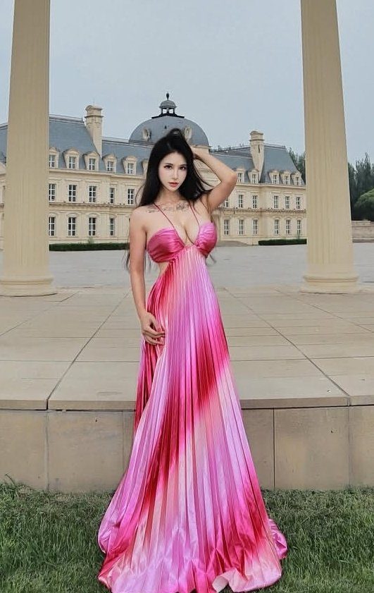 A woman in a flowing pink and red pleated dress poses outdoors in front of a grand building.