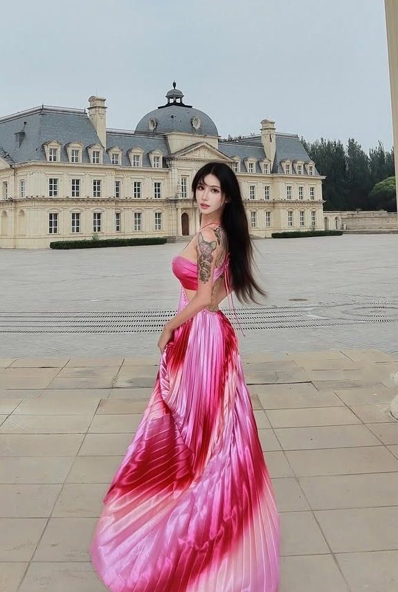 A woman with long black hair and a pink and red ombre dress stands outdoors in front of a large building.