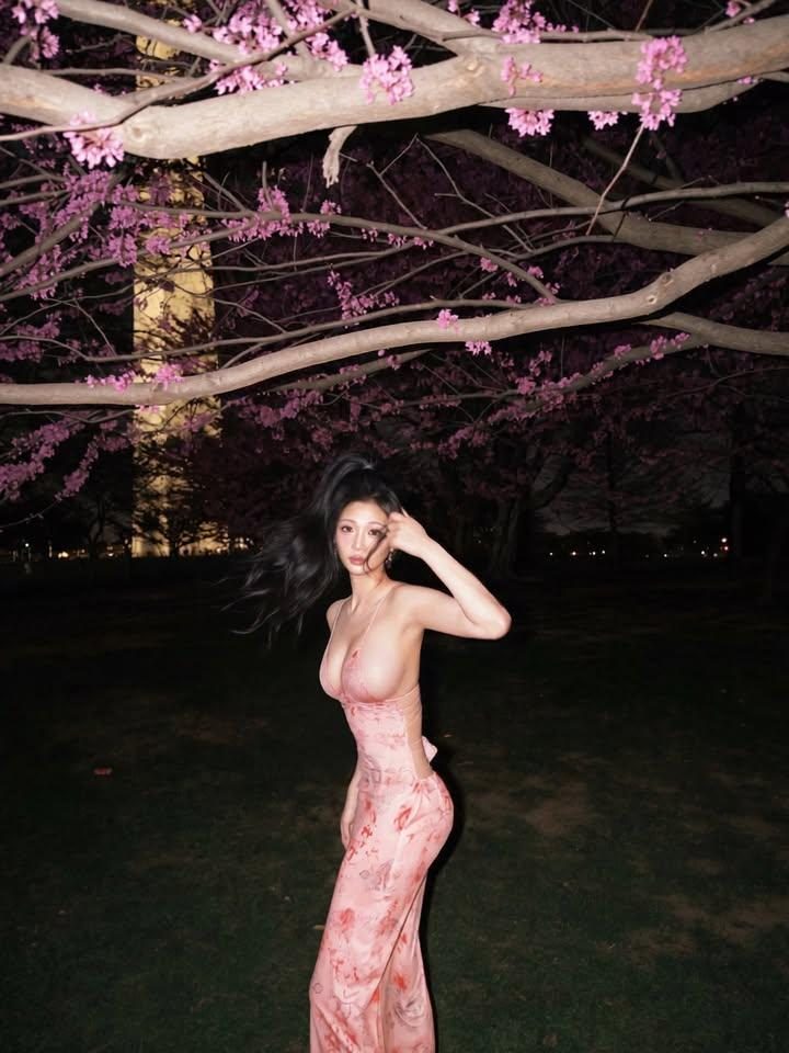 A woman in a pink floral dress stands outdoors at night under blooming trees.