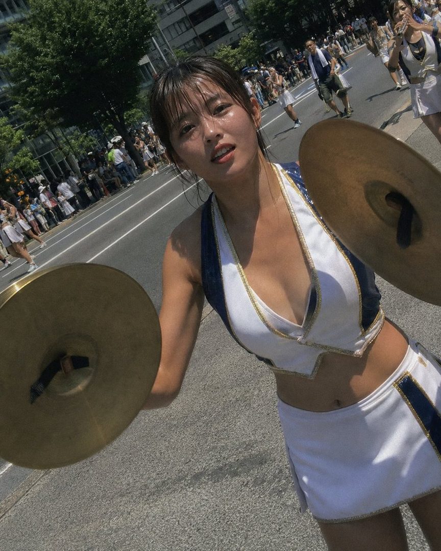 A young woman in a white and blue uniform with cymbals poses on a sunny street during a parade.