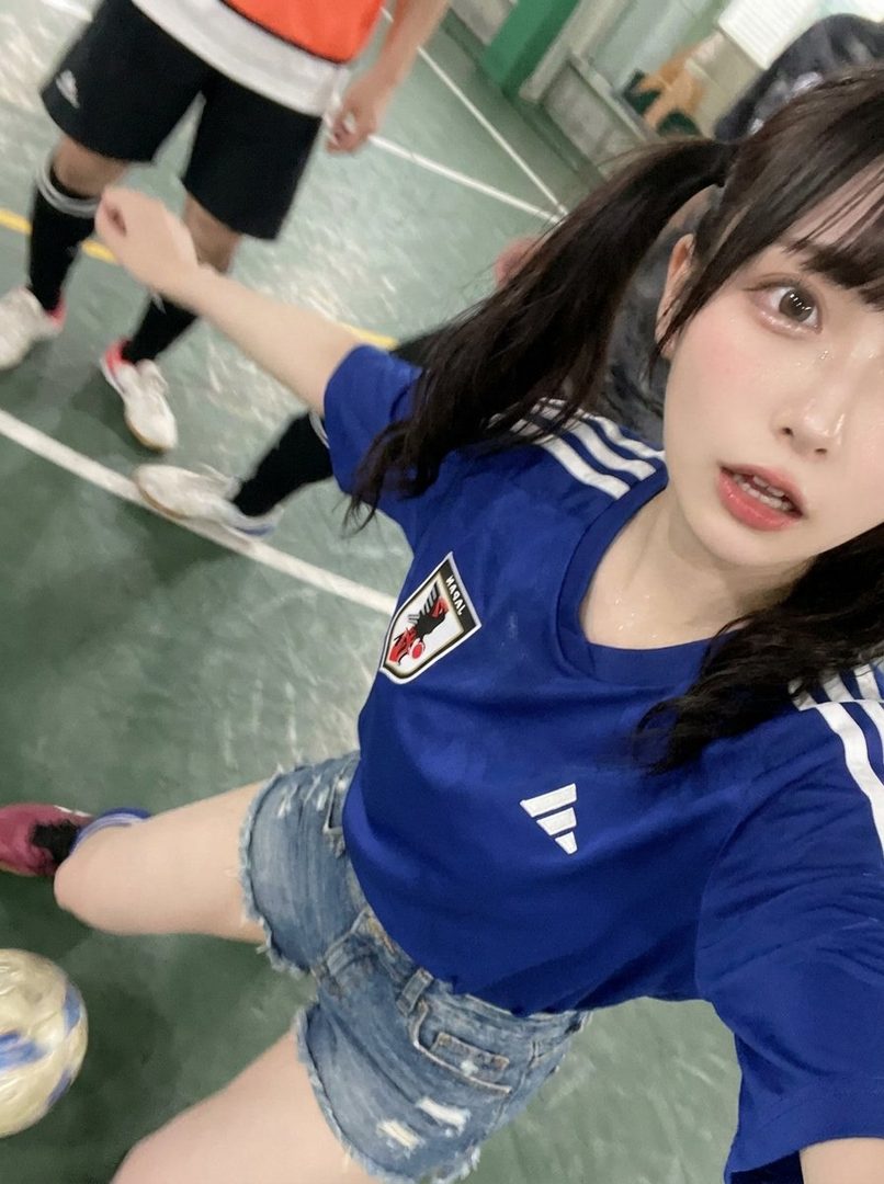 A young woman with black twintails and bangs, wearing a blue sports jersey and denim shorts, poses on an indoor court with a soccer ball nearby.