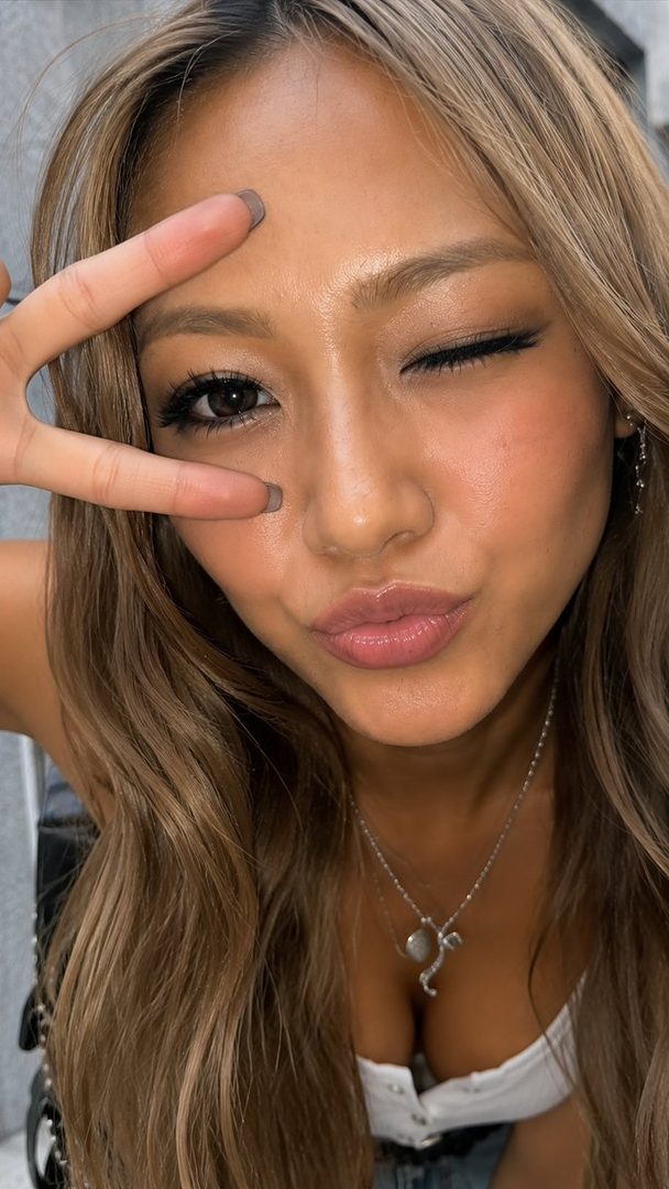 A young woman with long blonde hair winks at the camera while making a peace sign with her fingers.