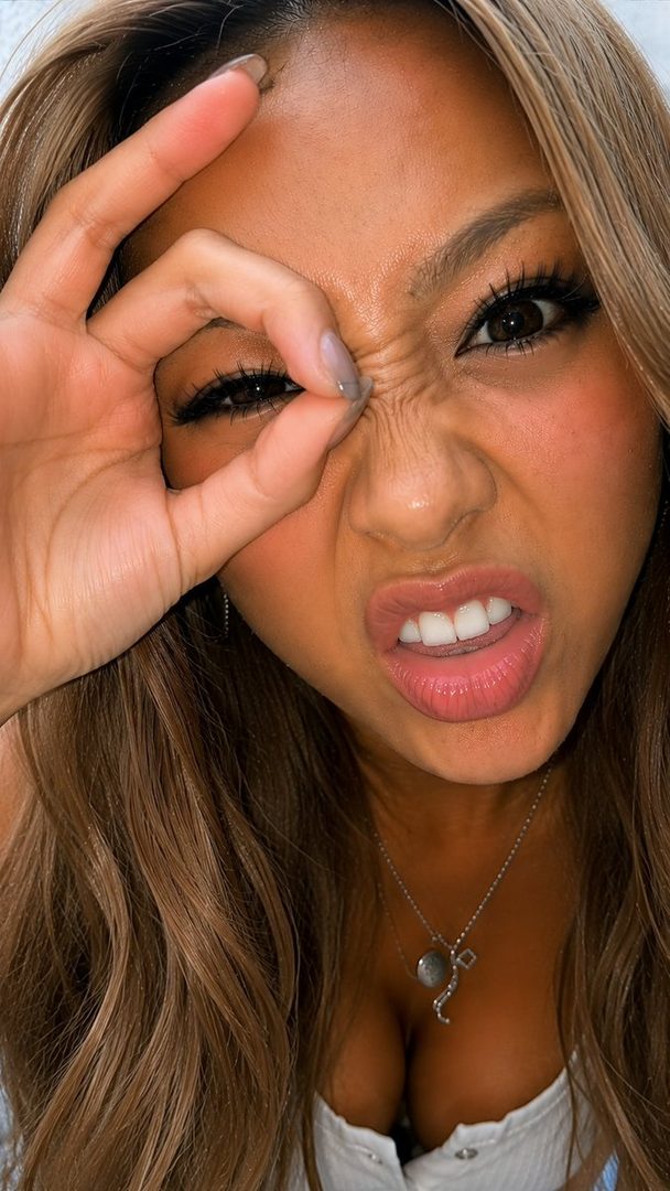 A woman with long brown hair makes a playful face, scrunching her nose and making an 'ok' sign with her hand over one eye.