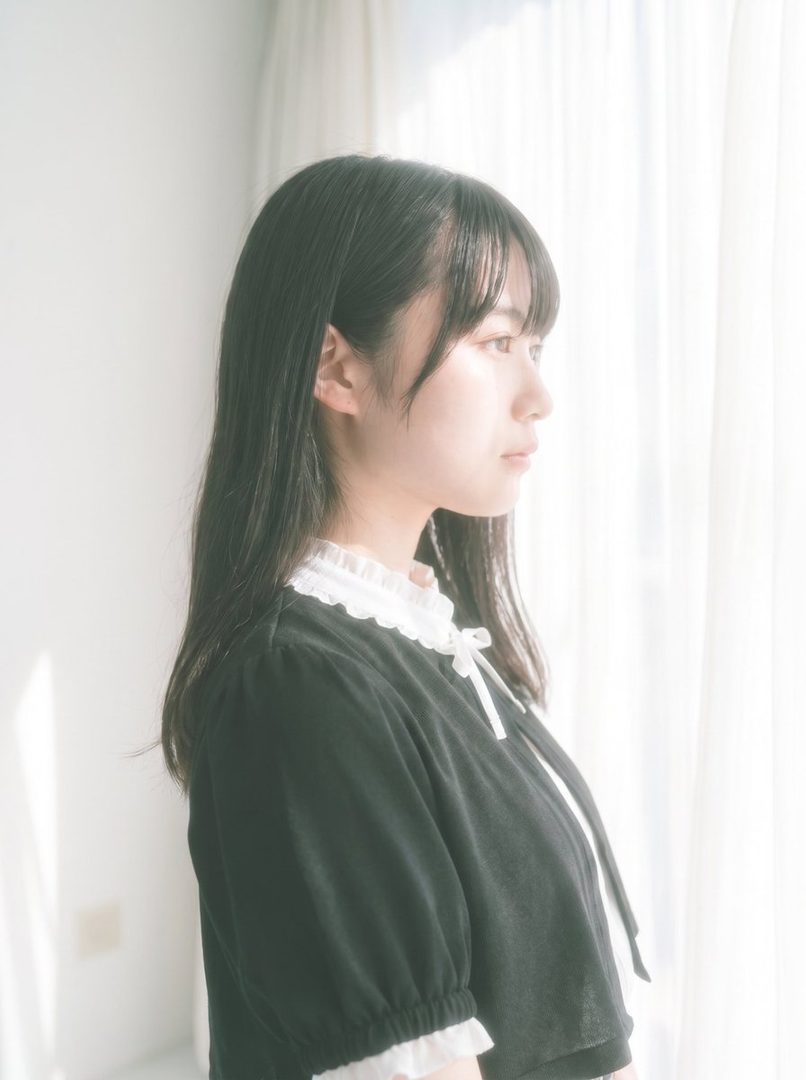 A young woman with long black hair and bangs, wearing a black blouse with a white ruffled collar, looks out a window with bright natural light.