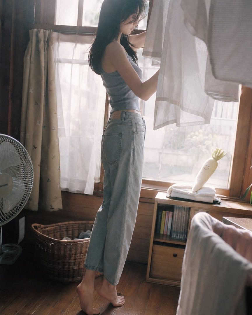 A young woman in a tank top and jeans stands barefoot by a window, hanging laundry indoors.