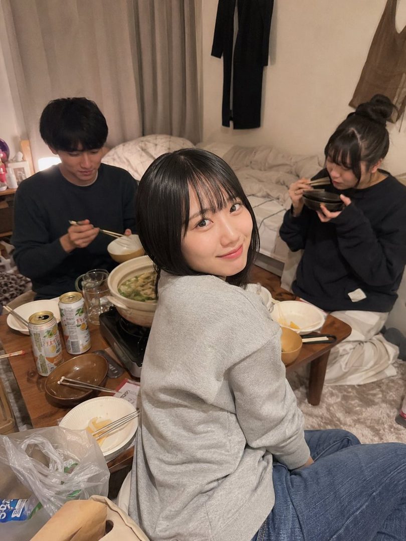 A young woman with short black hair and bangs smiles at the camera while sitting on the floor with two other people, eating from a hot pot.
