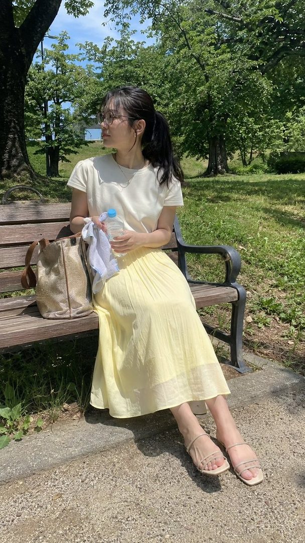 A young woman with black hair in a ponytail, wearing glasses, a white t-shirt, and a yellow skirt, sits on a park bench with a water bottle.
