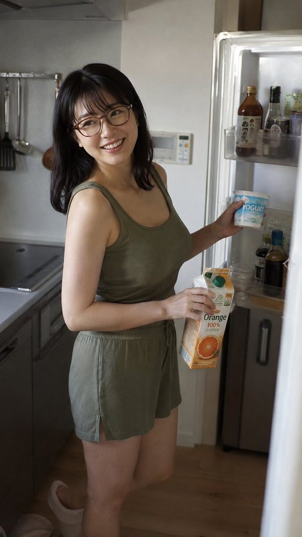 A smiling woman in glasses and casual wear stands in a kitchen, holding orange juice and yogurt from an open refrigerator.