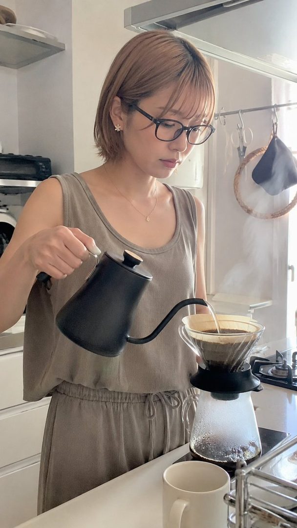 A woman with short blonde hair and glasses is making pour-over coffee in a bright kitchen.