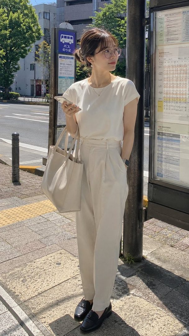 A young woman with glasses and a casual outfit stands at a bus stop on a sunny day, holding her phone and a tote bag.