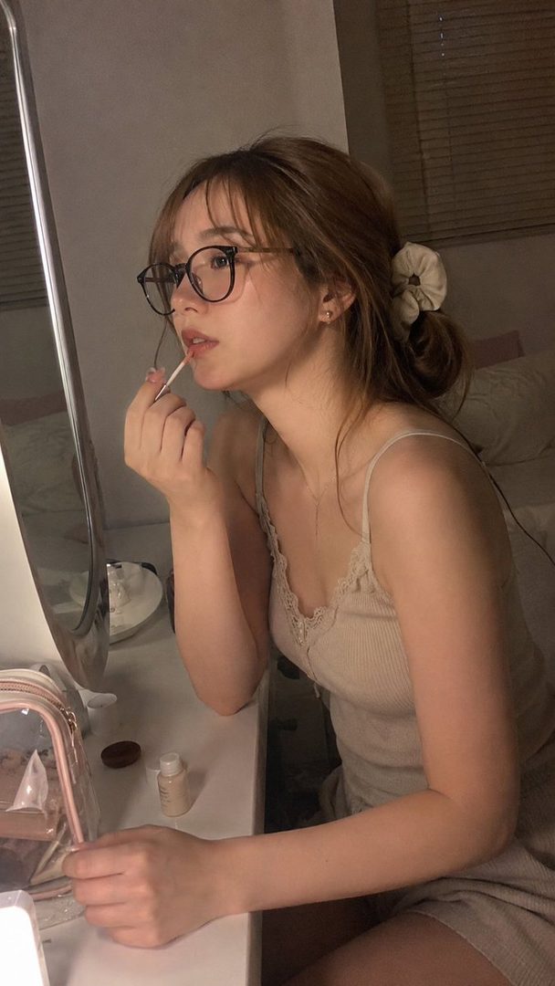 A young woman with glasses applies lip gloss while looking into a mirror at night.