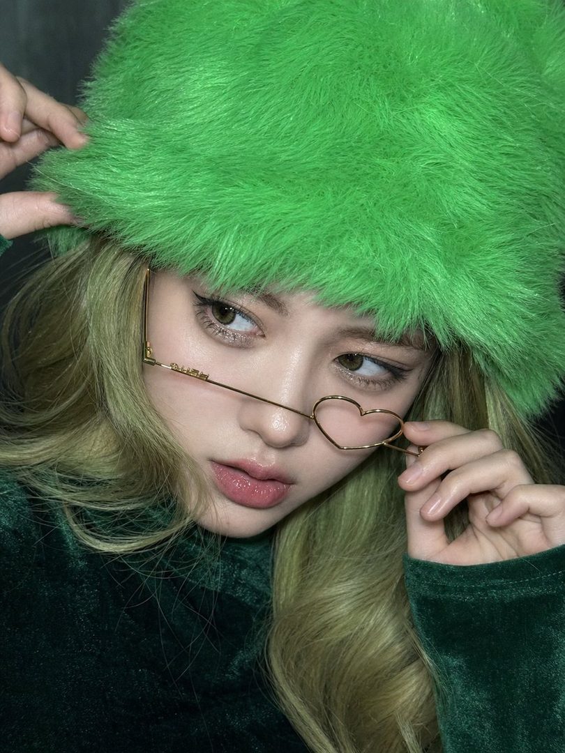 A woman with blonde hair and a bright green fuzzy hat looks away while adjusting heart-shaped glasses.