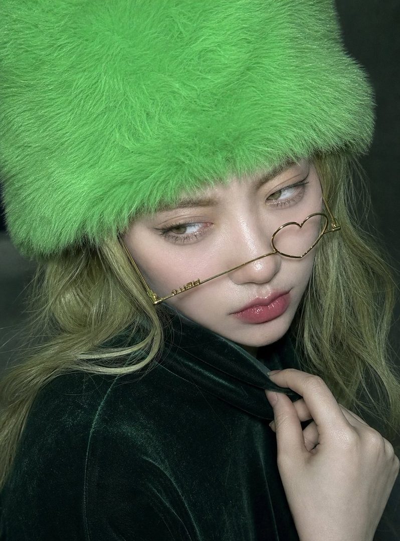 A woman with green hair and a bright green furry hat wears heart-shaped glasses and a dark green velvet top, looking away from the camera.