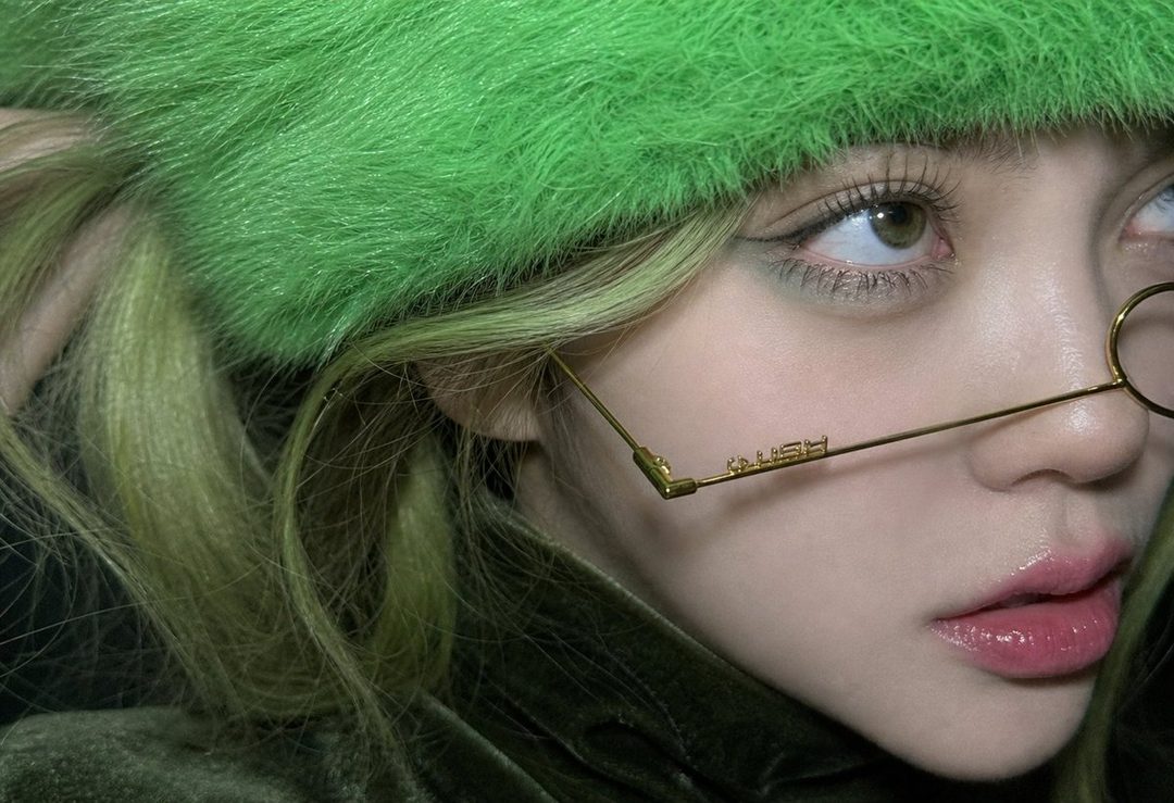A close-up portrait of a person wearing a bright green fuzzy hat and thin gold-rimmed glasses, with light green hair and glossy pink lips.