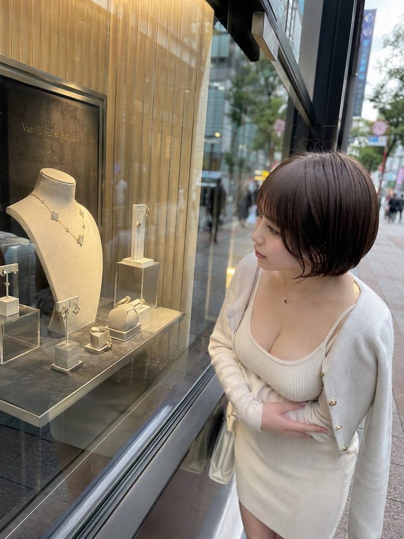 A young woman with short brown hair wearing a light-colored knit dress and cardigan looks into a jewelry store window on a city street.