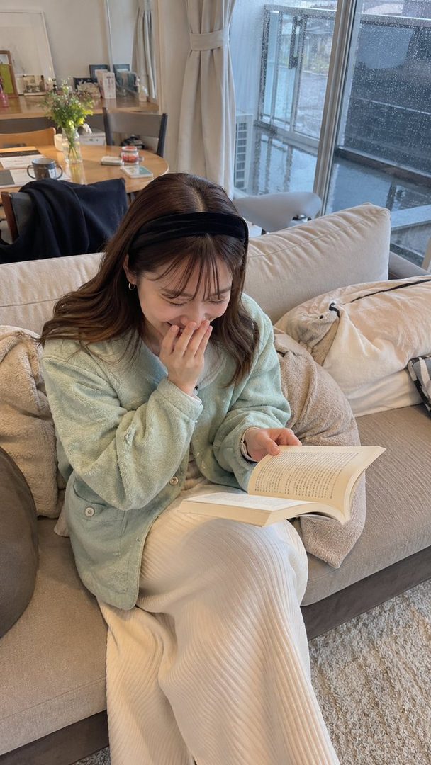 A young woman with long brown hair and bangs, wearing a light green cardigan and white pants, sits on a sofa reading a book and laughing.