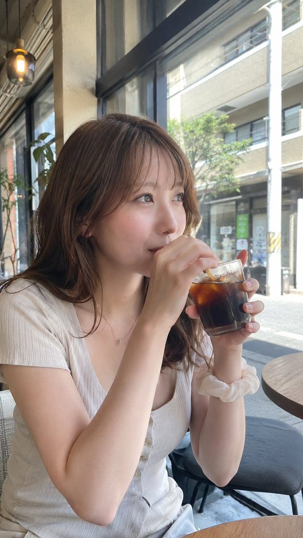 A young woman with brown hair sips iced coffee at a cafe, looking out the window.