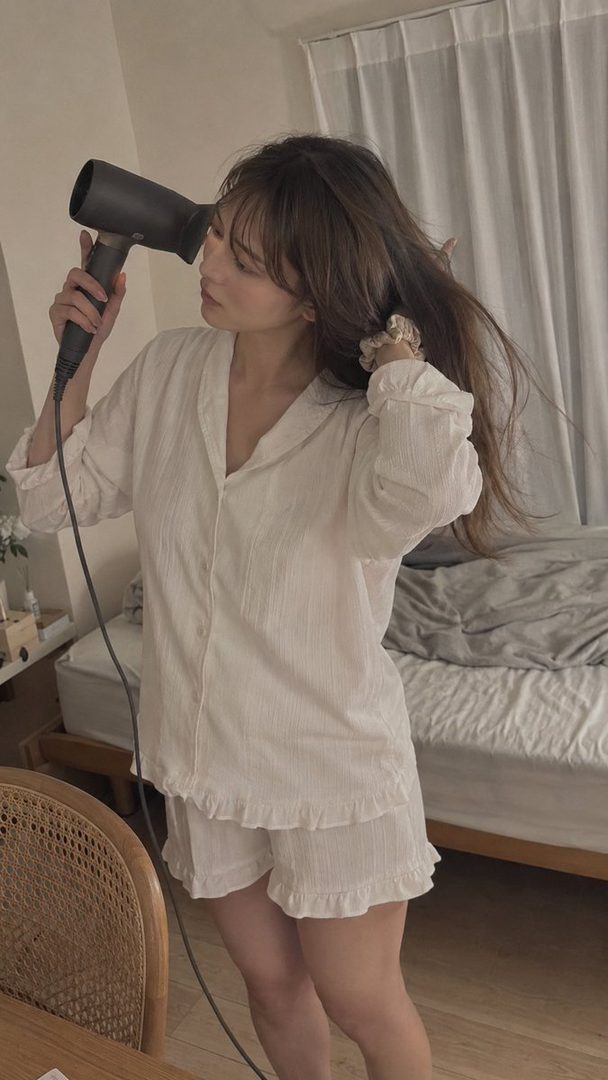 A young woman in white pajamas is drying her long brown hair with a hairdryer in a bedroom.