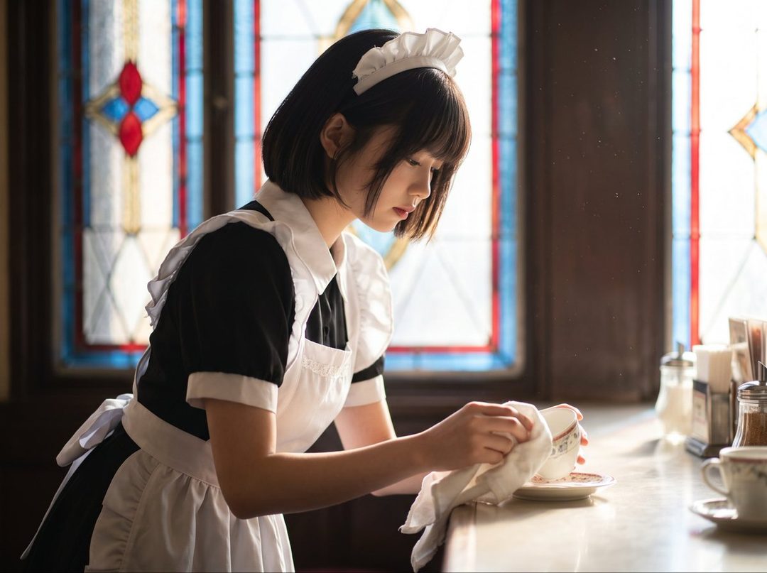 A young woman with short black hair and bangs, dressed in a maid uniform, is wiping a teacup in an indoor setting with stained glass windows in the background.