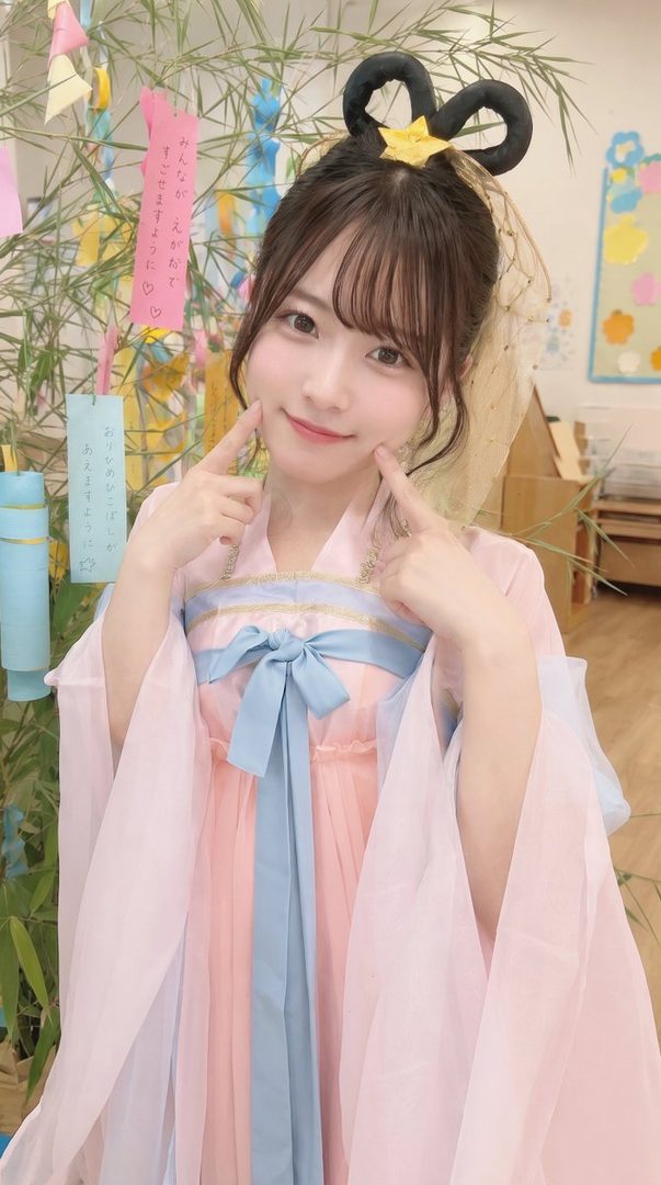 A cute Japanese woman in a pink and blue kimono-style outfit with a unique hair bun poses indoors.