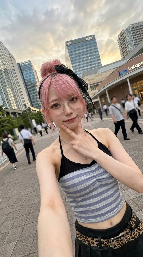 A young woman with pink hair and glasses takes a selfie on a city street, wearing a striped halter top and a black skirt with a leopard print belt.