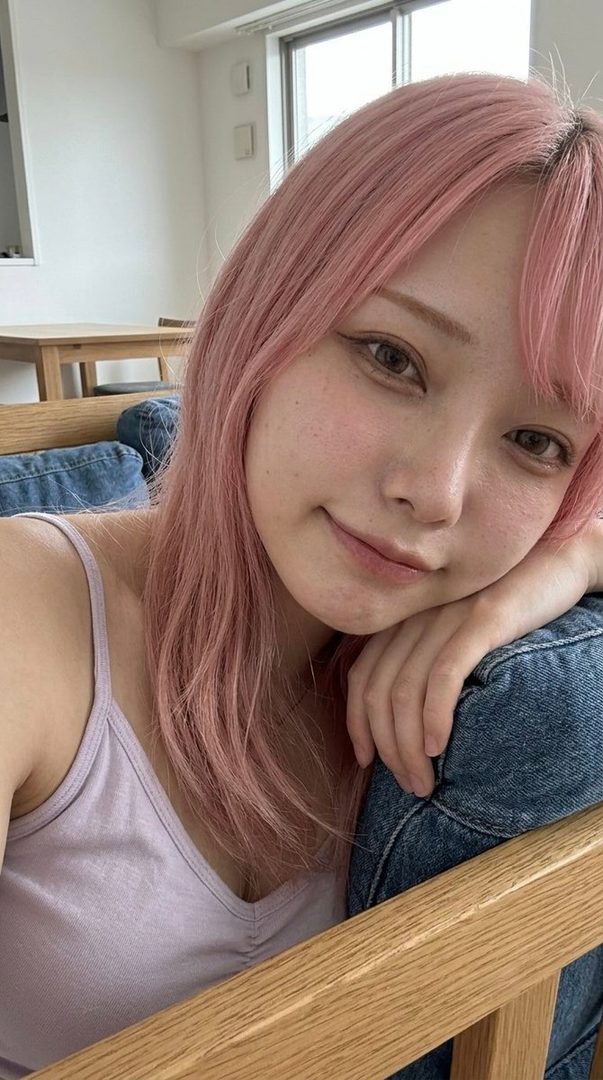 A young woman with pink hair takes a selfie indoors, leaning on a wooden chair with a gentle smile.