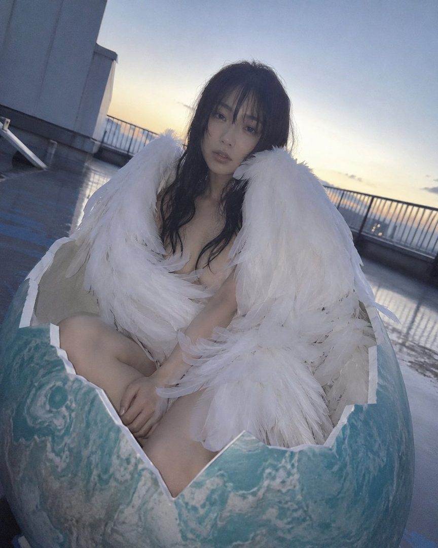 A woman with long black hair and bangs, wearing large white feathered wings, sits inside a broken eggshell on a rooftop at golden hour.