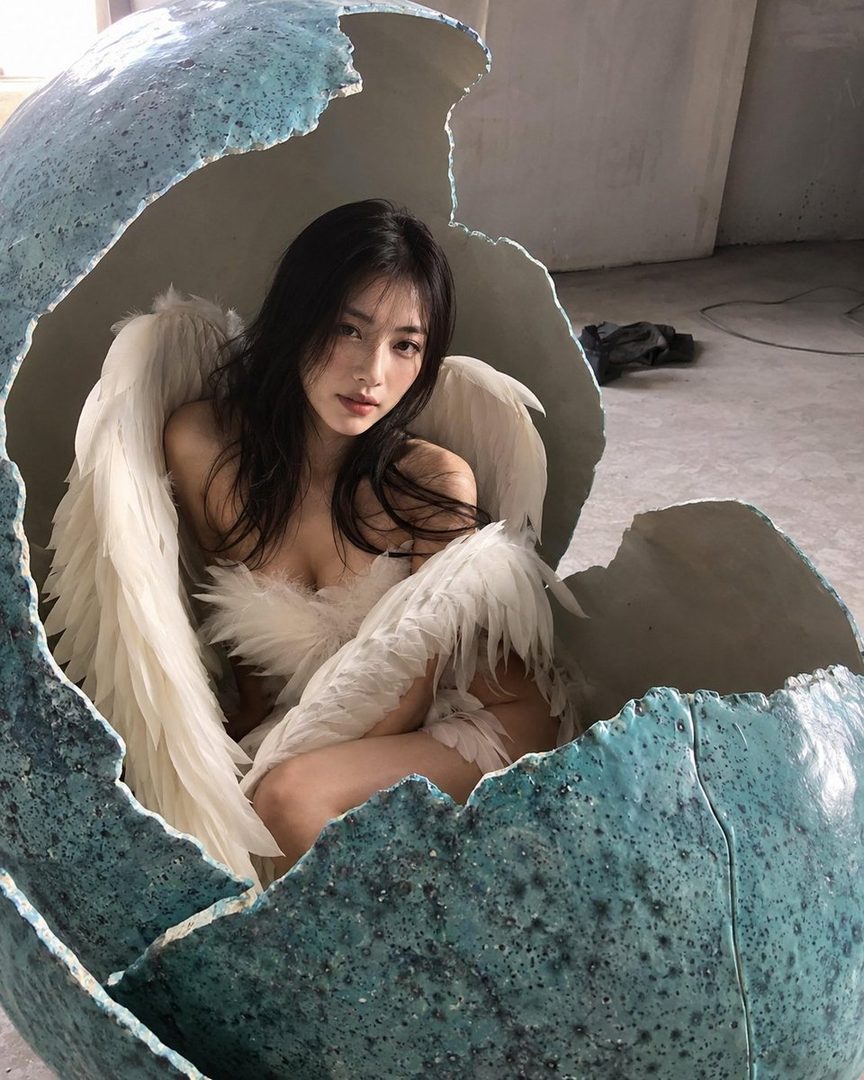 A woman with long black hair and white feathered wings sits inside a large broken eggshell.