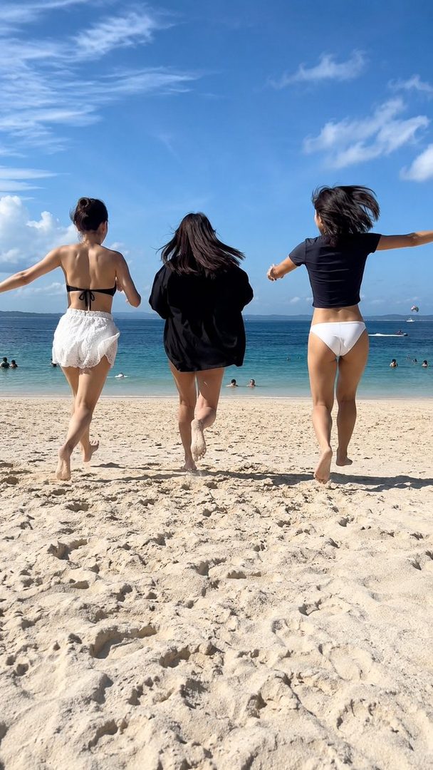 Three women in swimwear run towards the ocean on a sunny beach.