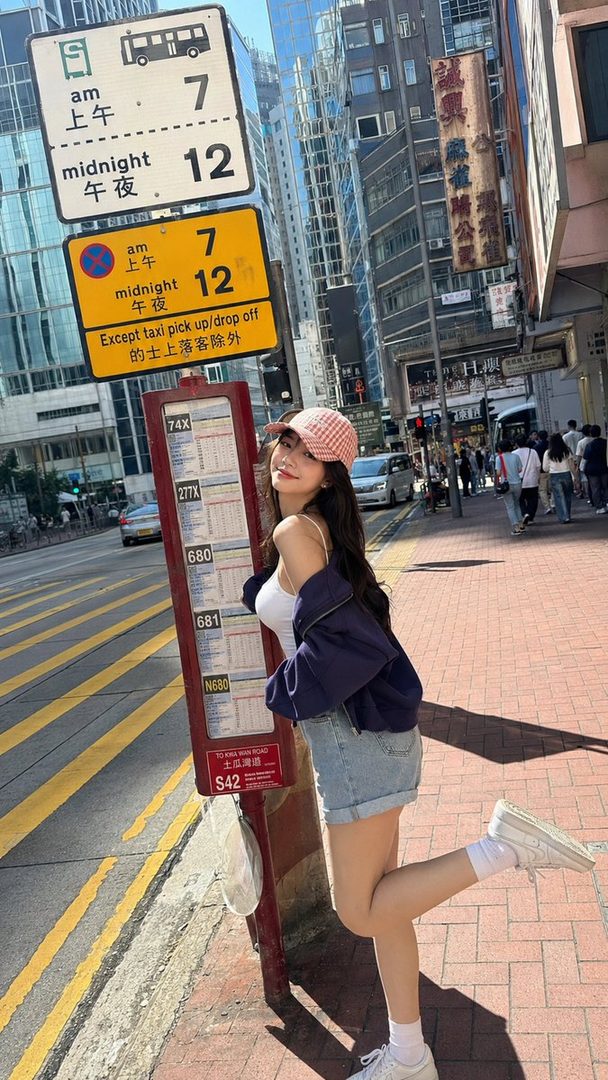 A young woman in a pink checkered cap, white tank top, and denim shorts poses playfully by a bus stop sign in a bustling city street.