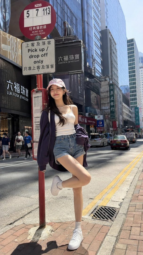 A young woman in a cap, white camisole, denim shorts, and purple jacket poses playfully by a bus stop sign on a sunny city street.